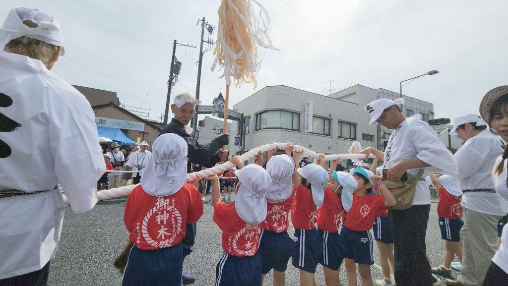 赤い「御神木祭」と背中に書かれたTシャツを着た、小さな子供たちがロープを持ち上げている様子